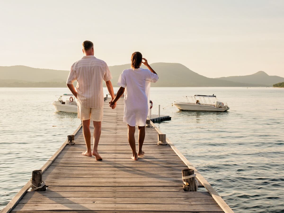 A man and a woman standing on a dock by a lake, holding hands. 