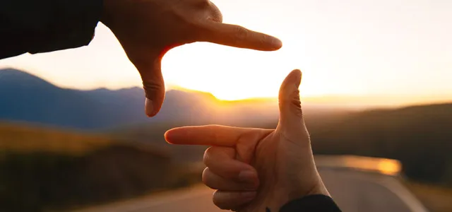 Two hands forming a rectangular frame around a sunset over a winding road and mountains.